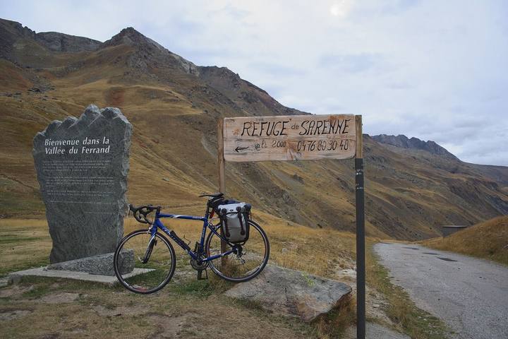Vélo chargé d'une sacoche devant la stèle de la vallée du Ferrand et le panneau du refuge, au col de Sarenne