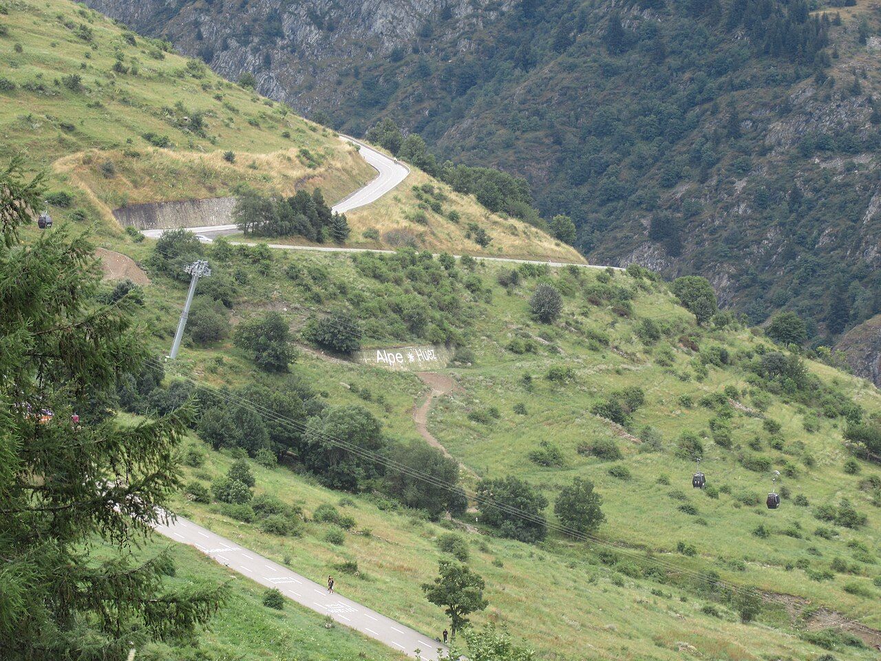 Haut de la montée : la route en lacets, l'inscription Alpe d'Huez sur le talus et les cabines de la télécabine