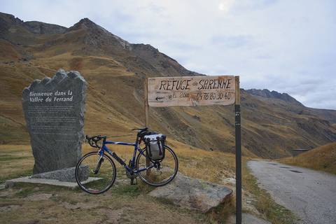 Vélo chargé d'une sacoche devant la stèle de la vallée du Ferrand et le panneau du refuge, au col de Sarenne